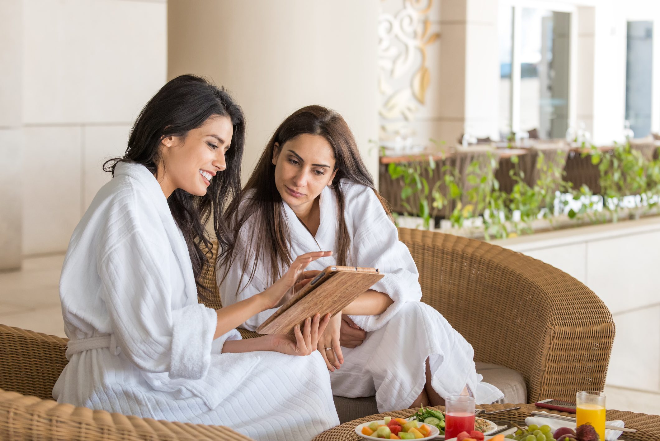 iStockalypse Dubai.  Arabian and mixed ethnic girlfriends relaxing in white bathrobes and eating a healthy spa lunch outside at a resort hotel while looking at a computer tablet.  Wicker furniture, with hotel cafe and landscaping in the background.  Dubai, UAE.